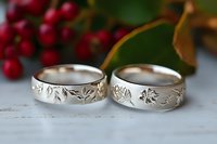 Silver wedding rings with engraved floral pattern on white wooden surface with red berries and green leaves in background, romantic jewelry photography.