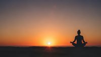 Woman silhouette meditating in lotus pose during golden sunset over ocean horizon. Peaceful yoga practice at beach for wellness and mindfulness concept.
