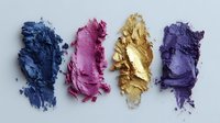 Crushed makeup eyeshadow pigments in blue, pink, gold, and purple arranged in a row on white background for cosmetic product photography.