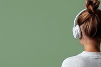 Young Asian woman with hair bun wearing white wireless headphones, viewed from behind against green background. Minimalist lifestyle concept.