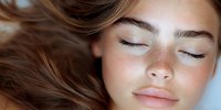 Close up portrait of young woman with natural makeup, closed eyes, and glowing skin lying down with brown hair spread out. Wellness and skincare concept.