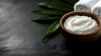 Creamy white moisturizing face cream in ceramic bowl with green tropical leaves and spa towels on dark stone background. Natural organic skincare concept.