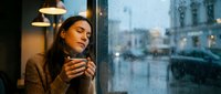 Young Caucasian woman holding coffee cup by rainy window in cozy cafe, eyes closed, enjoying warmth and calm atmosphere on a grey day.