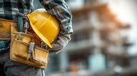 Construction worker holding yellow hard hat with tool belt at building site ready for safety work and industrial projects.