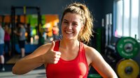 Smiling young Caucasian woman in red sports top giving thumbs up in gym, expressing positivity and fitness motivation.