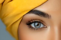 Close up of bright blue eye with long eyelashes and perfectly shaped eyebrow, wearing vibrant yellow headscarf against soft gray background. Macro beauty portrait.