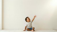 Young toddler with dark hair and medium skin tone sitting on floor with arm raised upward in joyful gesture, wearing striped shirt and denim shorts.