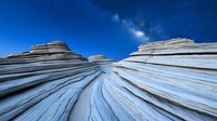 Dramatic layered white sandstone formations under starry night sky with Milky Way galaxy. Natural erosion patterns create flowing lines leading to cosmic view.