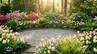 Circular stone patio surrounded by colorful spring flower garden with tulips, daffodils and blooming trees in golden sunlight for landscape design.