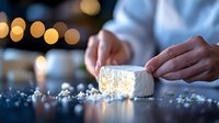 Soft marshmallow cube dusted with powdered sugar on dark surface with bokeh lights in background, close-up view of hands preparing sweet treat.