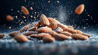 Raw almonds scattered on dark surface with powdered sugar explosion creating dramatic food photography effect against black background with floating nuts.
