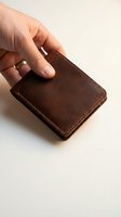 Hand holding a vintage brown leather bifold wallet with stitching detail on a light background, showcasing craftsmanship and style.