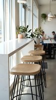 Modern cafe interior with wooden bar stools lined along white counter by large windows, potted plants, pendant lights, and blurred customers dining in background.