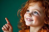 Happy redhead child girl pointing finger up with joyful smile against green background showing excitement and positive emotions for educational content.