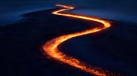 Glowing lava flow curves across dark volcanic landscape at night, creating bright orange and yellow molten stream for geological and nature content.