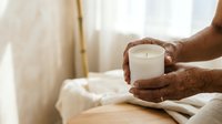 Elderly person with dark skin holding lit candle in white cup, seated indoors near window with natural light and neutral bedding, conveying warmth, comfort, mindfulness and peaceful moments.
