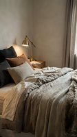 Cozy bedroom interior with unmade bed featuring neutral bedding, decorative pillows, brass reading lamp on wooden nightstand, and soft natural light from window with curtains.