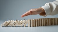 Female hand in white sleeve stopping wooden domino effect on light surface against gray background, representing crisis management and problem solving.
