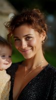Young mixed race woman with curly hair and natural makeup wearing black sparkly dress smiles warmly while holding toddler outdoors in soft natural lighting.
