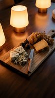 Wooden cutting board with assorted cheeses, grapes, and figs arranged under warm ambient lighting from brass table lamps, suitable for food styling and entertaining concepts.