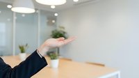 Businesswoman in dark blazer gestures with open palm in modern office conference room with wooden table, potted plants, and contemporary lighting fixtures.
