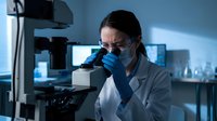 Female scientist in lab coat and blue gloves examining samples through microscope in modern research laboratory for medical analysis and scientific discovery.
