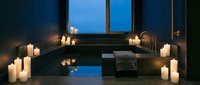Luxurious spa bathroom with sunken bathtub surrounded by lit candles, dark tiles, and panoramic windows overlooking twilight sky, creating serene wellness and relaxation ambiance.
