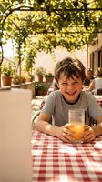 Happy young boy enjoying fresh orange juice at outdoor cafe terrace with vine pergola and checkered tablecloth during sunny summer day.