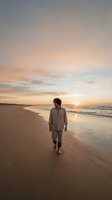Man with dark curly hair, appearing to be in his 30s of mixed ethnicity, walks barefoot along sandy beach at golden hour sunset. Wearing casual beige linen shirt and pants. Serene coastal scene with gentle waves and warm sky.
