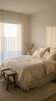 Serene bedroom with cream linen bedding, wooden bench, dried pampas grass in vase, sheer curtains filtering natural light through large window, minimalist interior design.