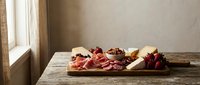 Rustic charcuterie board with cured meats, cheese and nuts on wooden table by window. Artisanal appetizer platter for entertaining and food photography.