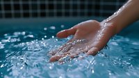 Hand touching clear blue swimming pool water with splashing droplets creating ripples and reflections in sunlight for relaxation and wellness concepts.