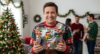 Middle-aged Caucasian man in colorful Christmas sweater holding champagne flute at holiday party with decorated tree and festive garland background.