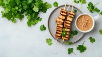 Grilled tofu skewers on gray plate with peanut dipping sauce, surrounded by fresh cilantro leaves on white background.