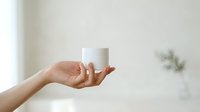 Woman's hand holding blank white cosmetic jar against neutral background with soft natural light and blurred greenery, suitable for skincare, beauty product packaging mockups.