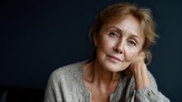 Elegant senior woman with blonde hair in gray sweater against dark background, contemplative expression with hand resting on neck.