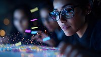 Young Asian woman in glasses illuminated by colorful digital light, looking at interactive display with glowing particles and confetti in dark environment.