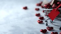 Mini shopping cart with red glitter hearts and blank gift tag on gray concrete background, romantic holiday shopping concept for Valentine's Day celebration.