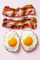 Crispy bacon strips and fried eggs isolated on pink background for breakfast menu design and culinary projects.
