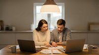 Young Caucasian couple working together at home office desk with laptops and documents under warm pendant lighting in modern kitchen setting.