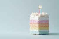 Colorful rainbow layer cake slice with white frosting and lit birthday candles on pastel blue background. Minimal party celebration concept with copy space.