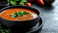 Fresh tomato soup in black bowl garnished with herbs and spices on dark stone surface. Healthy comfort food for restaurant menus and cooking blogs.