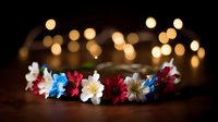 Delicate floral crown with red, white and blue flowers on dark wooden surface against blurred warm bokeh lights background creates romantic atmosphere.