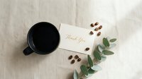 Black coffee mug beside a thank you card, scattered coffee beans, and eucalyptus branch on linen, flat lay, minimal style.