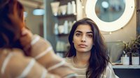 Young Middle Eastern woman with long dark hair in beige sweater sitting at beauty salon with ring light and mirror reflection, fashion and beauty concept.