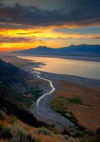 Dramatic sunset over mountain lake with winding river through valley, golden light illuminating clouds while silhouetted peaks frame tranquil waters and meandering shoreline.