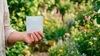 Caucasian person holding blank white card in hand against blooming garden background with colorful flowers for greeting, invitation, or promotional messaging.
