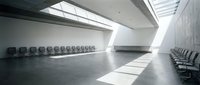 Modern minimalist waiting area with rows of black chairs in bright white interior space with natural lighting from skylights and geometric architecture.