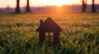 Small wooden house model on green grass field at golden sunset with warm light streaming through windows for real estate and home concepts.