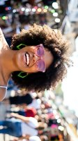Happy young African American woman with curly hair wearing pink sunglasses smiling joyfully at outdoor festival with colorful bokeh lights in background.